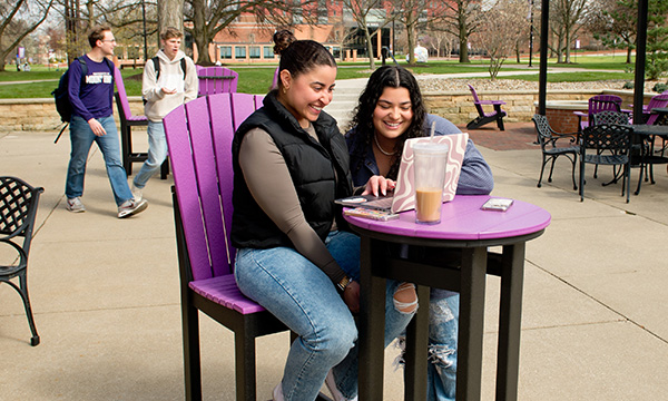 Two students hanging out on the Campus Grounds patio, viewing content on a laptop