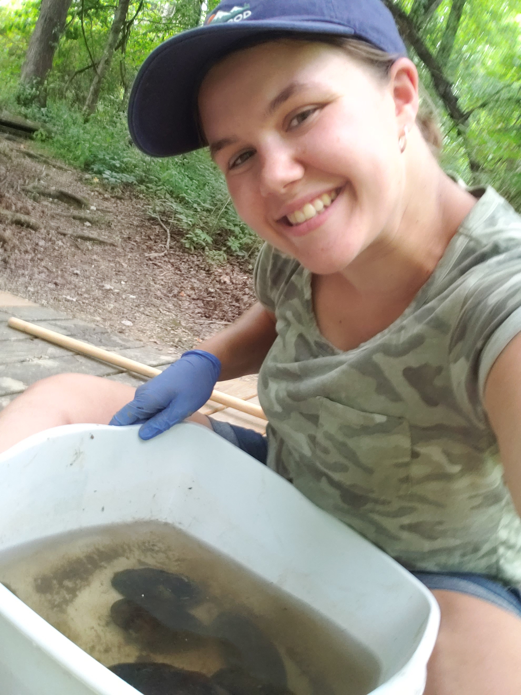 Emily Becker '23 poses in a hat in gloves at the Nature Center, holding a small tub of mollusks in water.