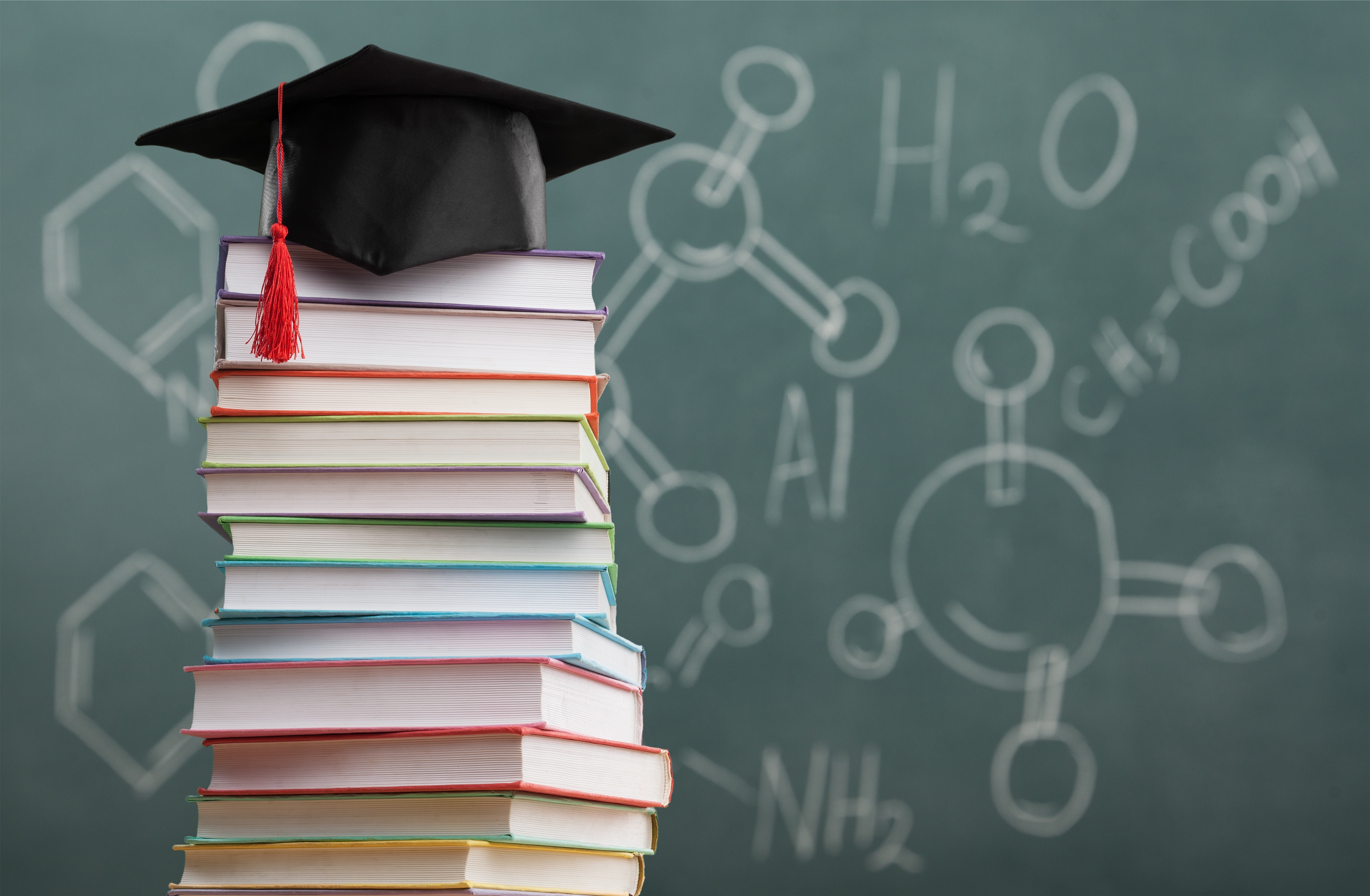 Photo of a chalk board with science formulations and a stack of books with a graduation cap on top