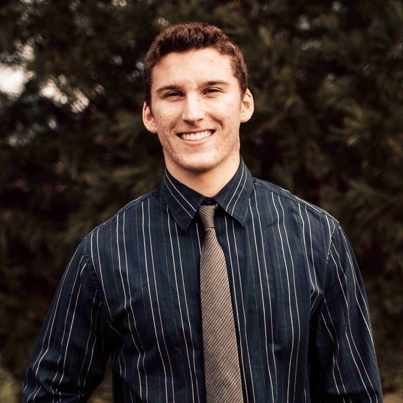 Samuel Atkinson stands smiling against a tree backdrop